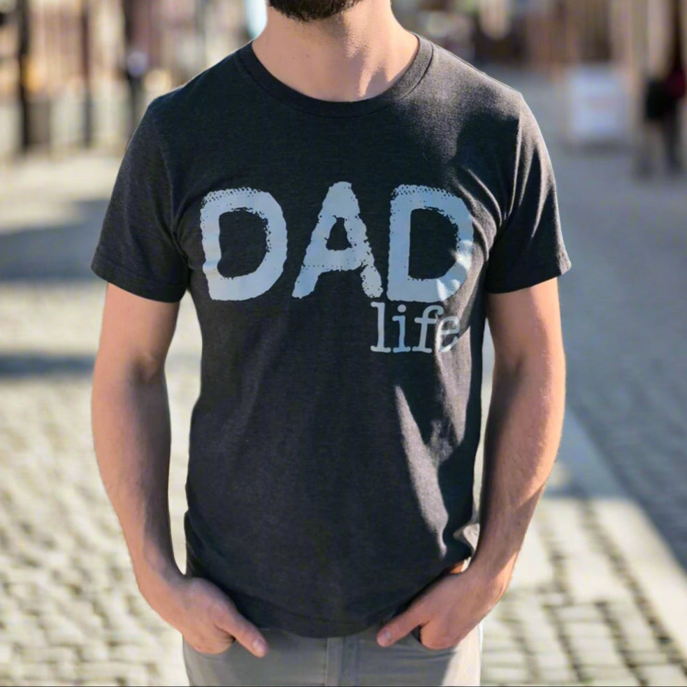 A bearded man stands outdoors on a cobblestone street wearing the Dad Life Tee, a dark shirt with DAD life in bold white letters; his hands are in his pockets and his face is partially out of frame.
