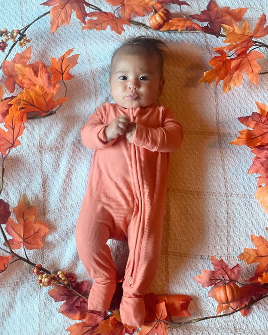 A baby in the Coral Ribbed Solid • Bamboo Zippy (Updated Fit) relaxes on a blanket, surrounded by autumn leaves and pumpkins—a festive fall scene. This gentle bamboo onesie is perfect for sensitive skin.