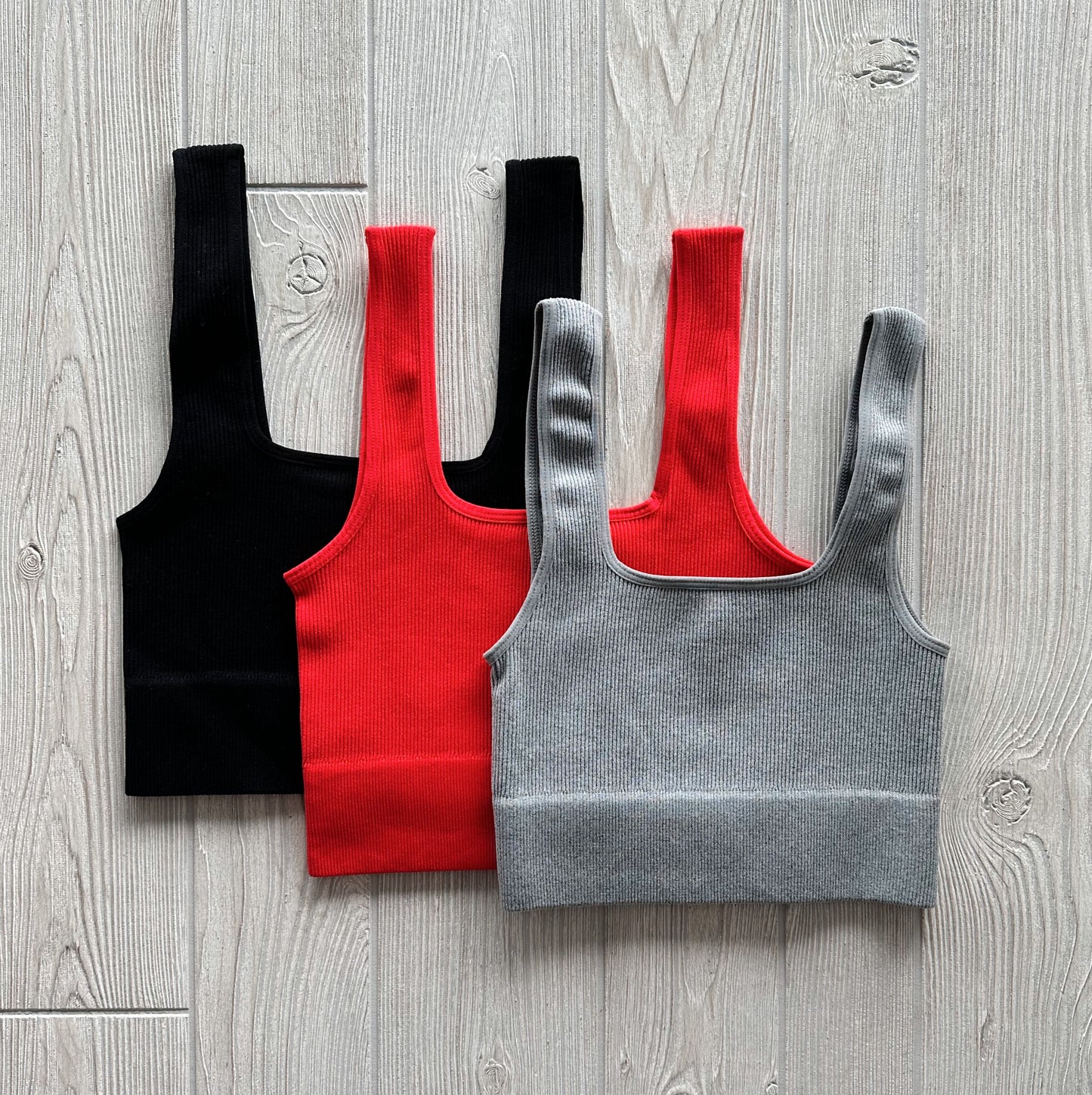 Three Iris Crop tank tops in black, red, and gray are arranged on a light wooden floor, overlapping to display their soft, ribbed, and stretchy fabric.