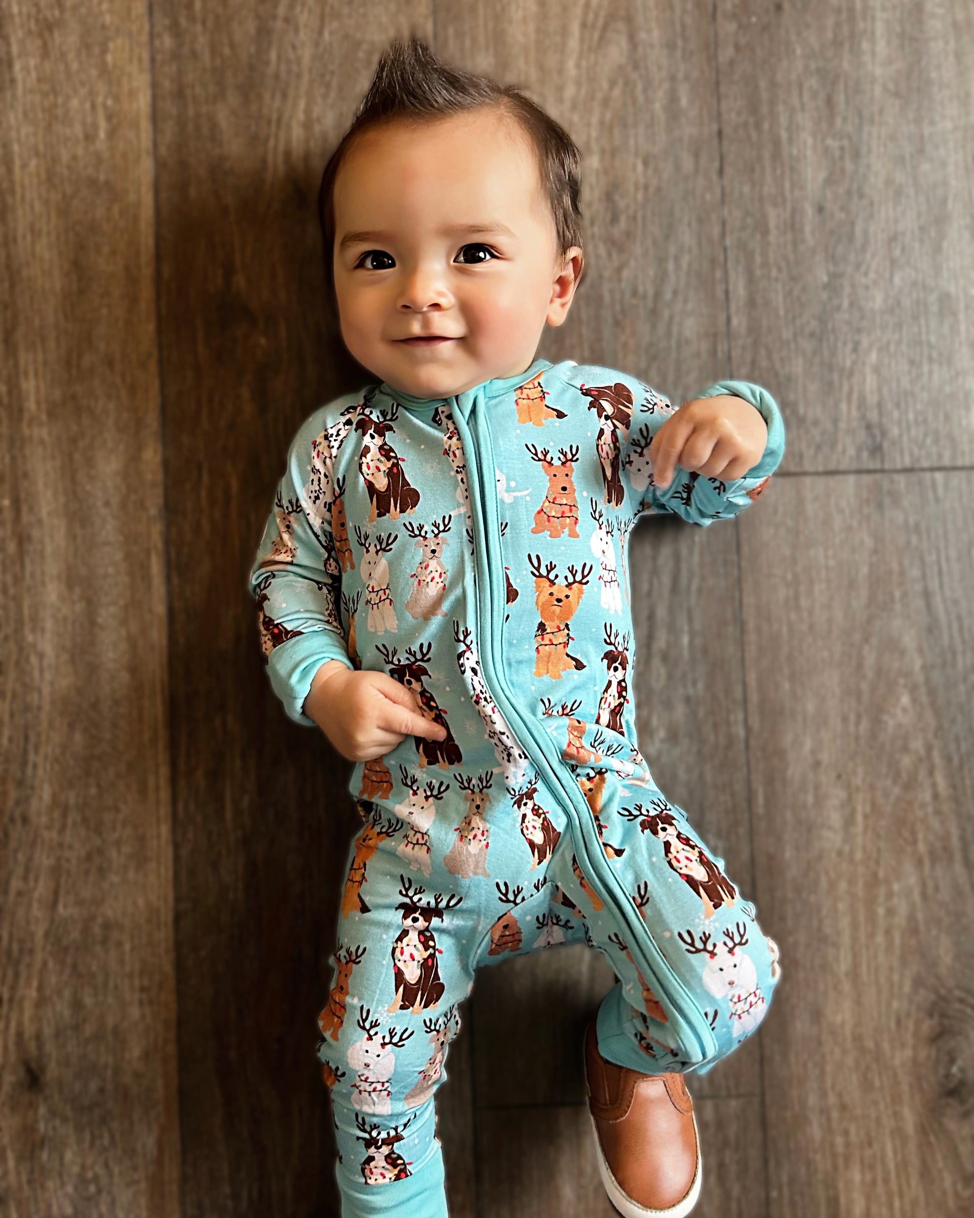 A smiling, dark-haired baby stands on a wooden floor wearing the Festive Friends • Bamboo Zippy (Updated Fit), featuring animal and antler patterns in blue. The baby has one hand raised and wears a single brown shoe.