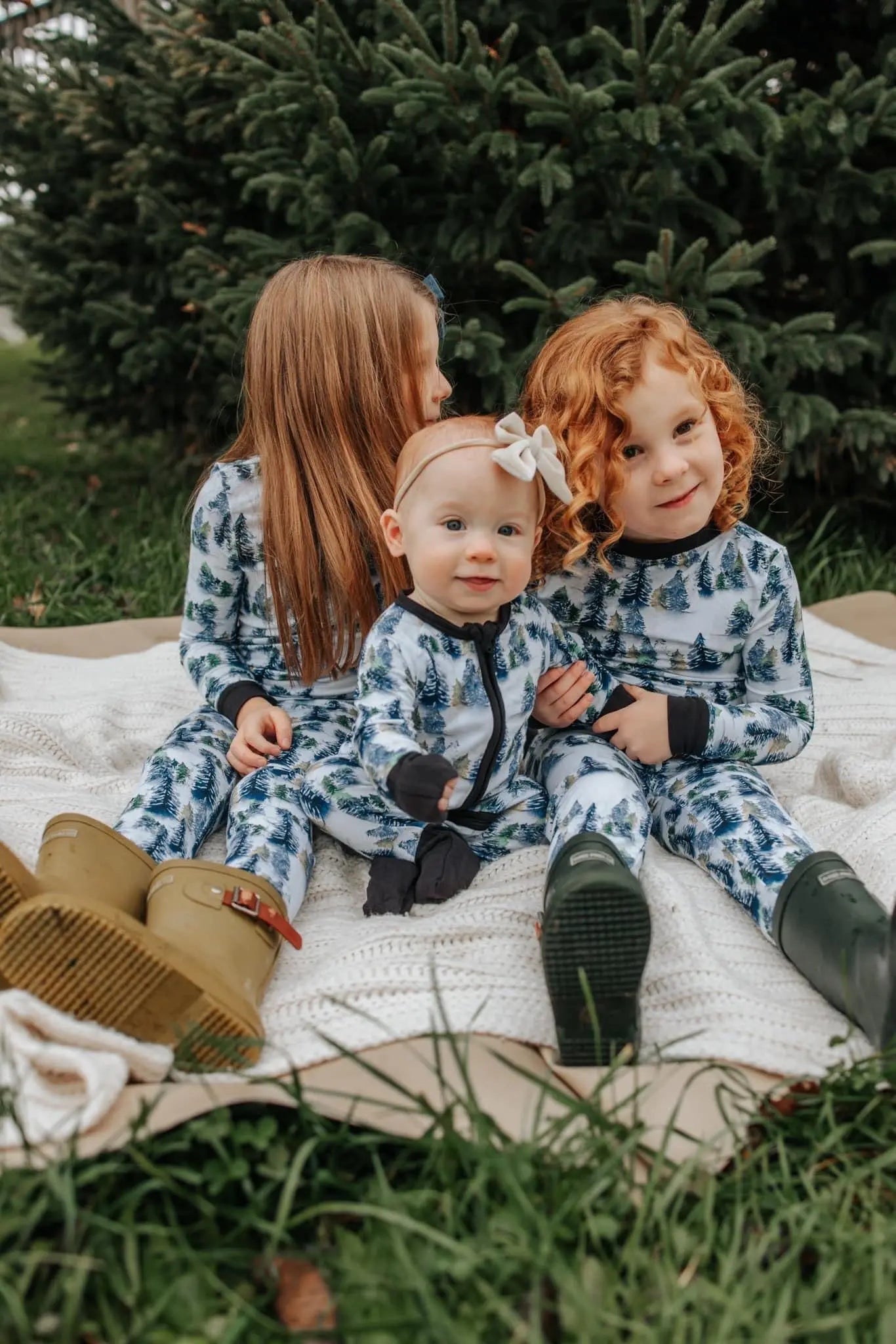 Three young children sit on a blanket outdoors by pine trees, wearing our Snowy Trees • Bamboo Zippy (Updated Fit) pajamas. One has red curls, another has straight blonde hair, and the baby in front wears a bow headband.