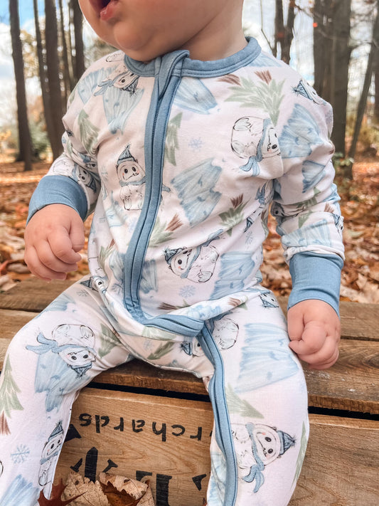 A baby in the Abominable Snowman • Bamboo Zippy (Updated Fit)—light blue and white pajamas with cute snowman designs—sits on a wooden crate outdoors, surrounded by autumn leaves and trees.