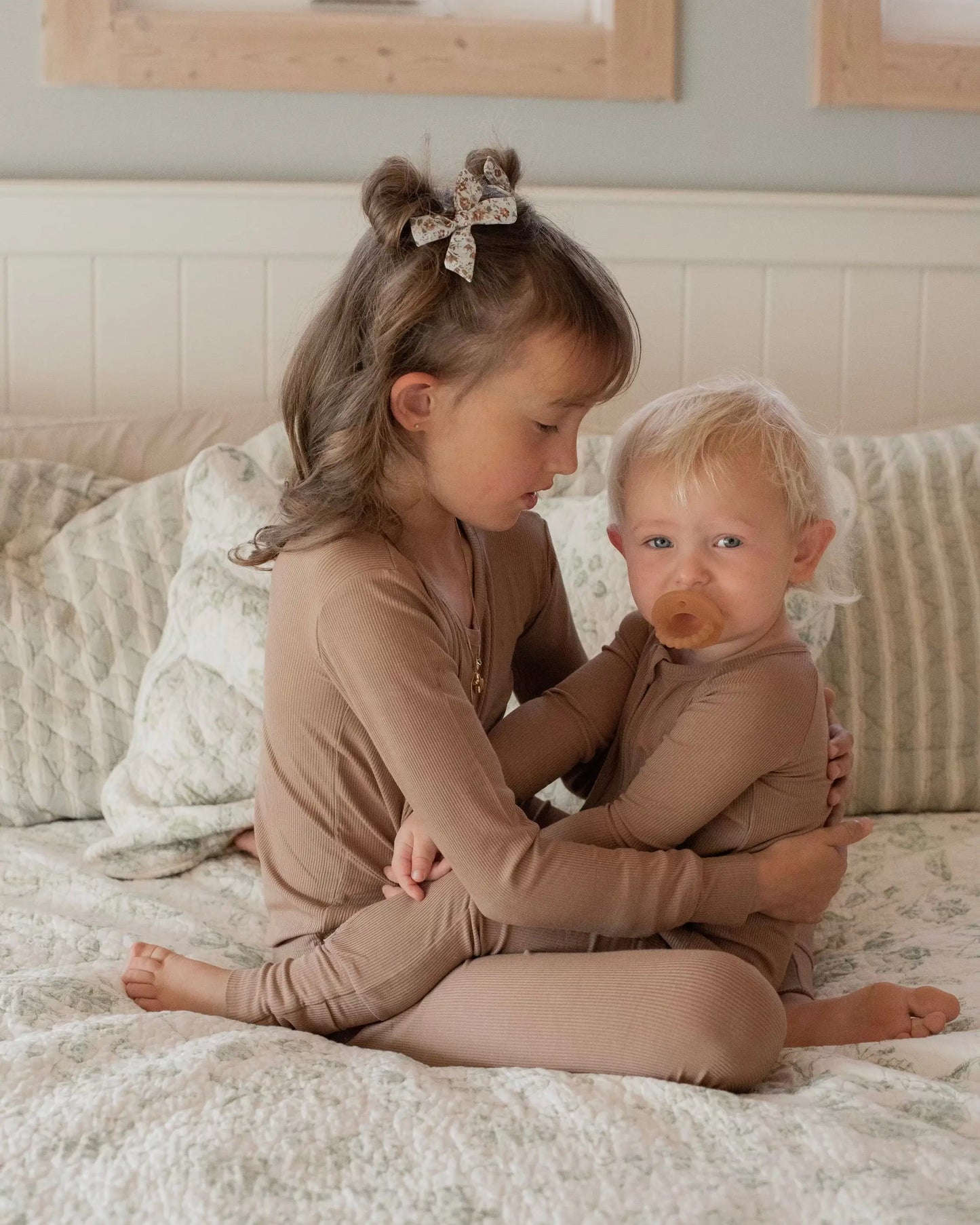 A young girl with a hair bow sits on a bed, hugging a toddler with a pacifier. Both wear matching Chestnut Ribbed Solid • 2 Piece Bamboo Sets—ultra-soft and ideal for sensitive skin. The bed has light, patterned bedding and pillows.