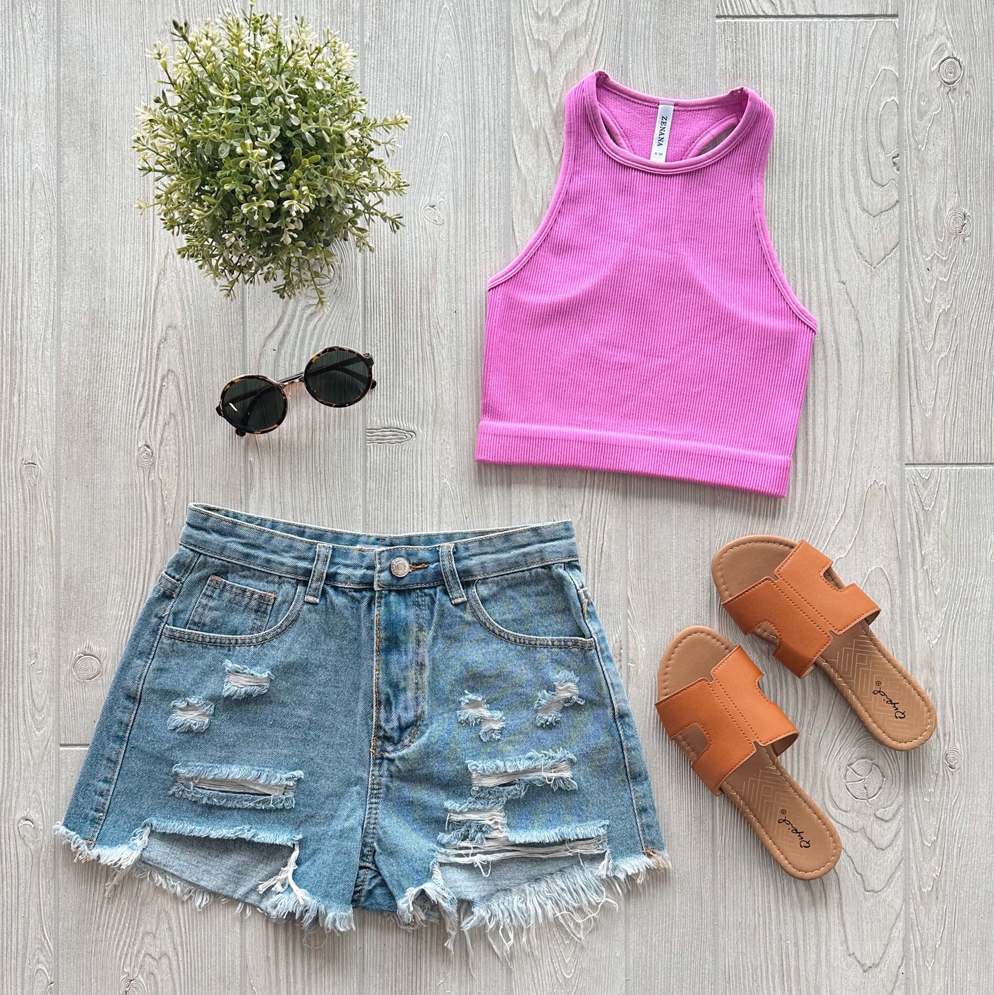Flat lay of a summer outfit: Sophia High-Neck Crop in orchid, distressed denim shorts, tan slip-on sandals, black round sunglasses, and a small potted plant on a light wooden background.