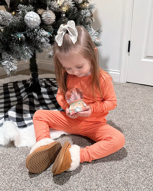 A young girl in our Coral Ribbed Solid • 2 Piece Bamboo Set (Updated Fit) sits on the carpet by a Christmas tree, smiling at a festive ornament—her bamboo viscose pajamas are gentle and perfect for sensitive skin.
