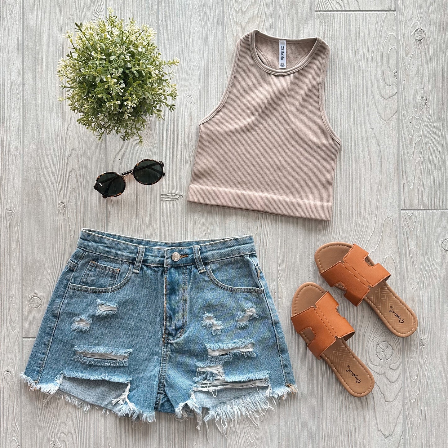 Flat lay of a casual summer outfit featuring the Sophia High-Neck Crop in Mocha, distressed denim shorts, tan slip-on sandals, round black sunglasses, and a small potted plant on a light wooden floor.