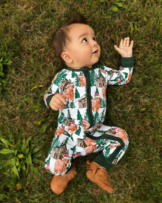 A baby in a Santa Cows • Bamboo Zippy (Updated Fit) onesie lies on green grass, reaching up—made from soft bamboo, its perfect for sensitive skin.