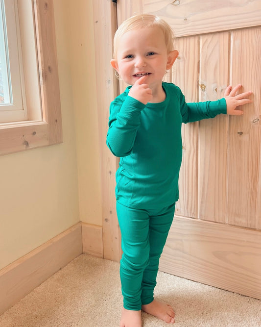A smiling toddler with short blond hair wears the Emerald Ribbed Solid • 2 Piece Bamboo Set (Updated Fit)—a green long-sleeve top and matching pants made for sensitive skin—standing on beige carpet by a wooden door, one hand on the door.