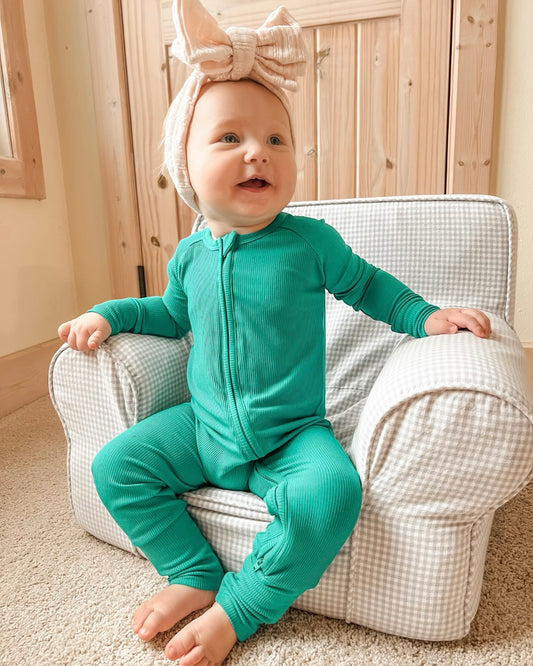 A smiling baby in the Emerald Ribbed Solid • Bamboo Zippy (Updated Fit) onesie and a large pink bow headband sits on a small checkered armchair in a cozy room with wooden doors and beige carpet.