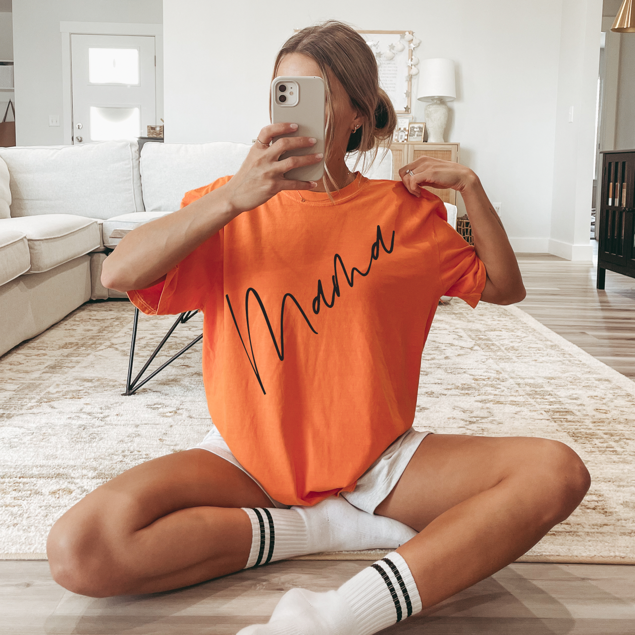 A woman sits cross-legged on a rug in her living room, wearing the Mama Diagonal Burnt Orange Dyed Tee and white shorts, taking a mirror selfie with her phone.
