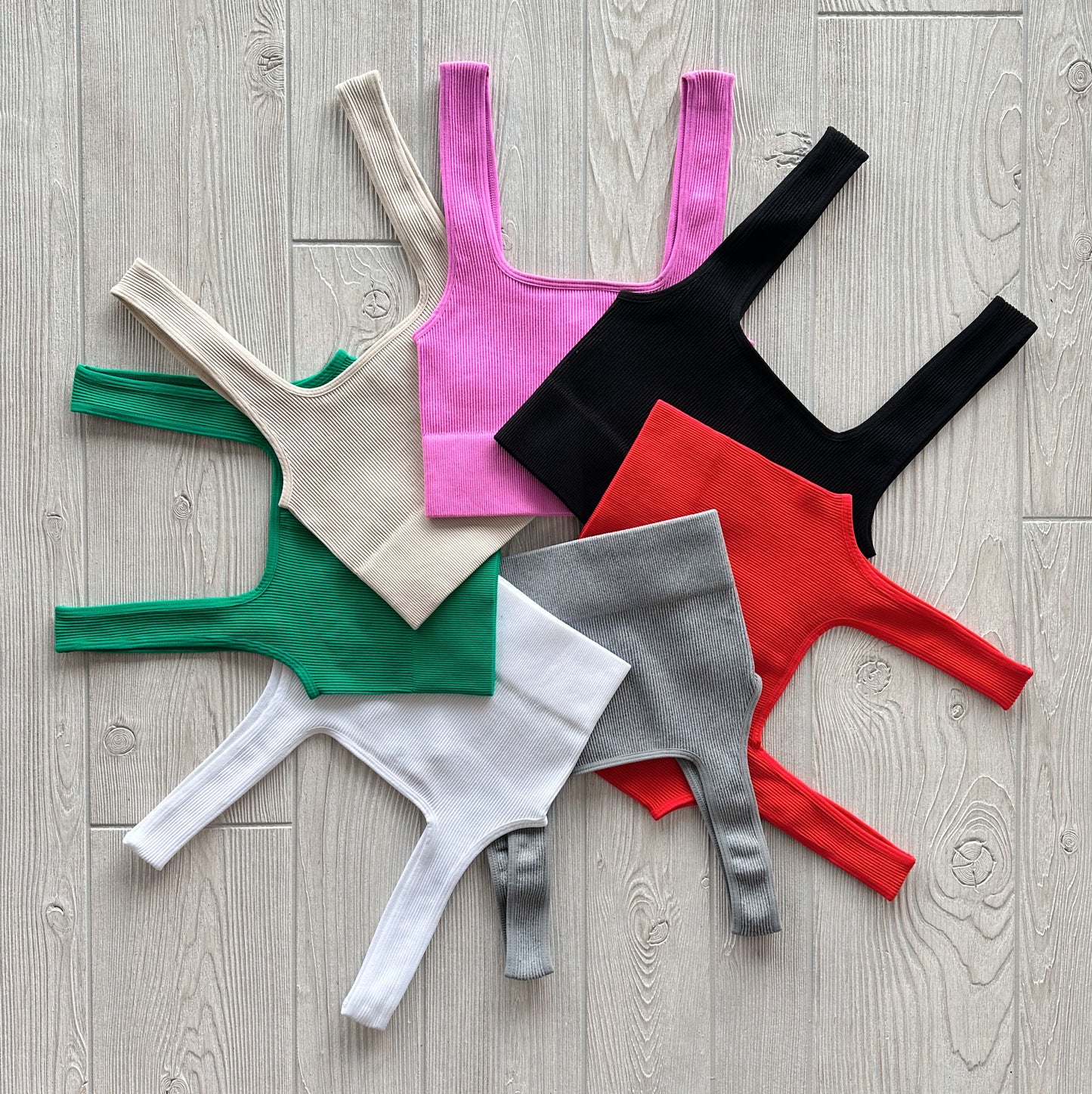 Six Iris Crop tops in beige, green, pink, black, red, and white form a starburst pattern on a light wood floor. Their ribbed fabric and outward straps create a striking display.