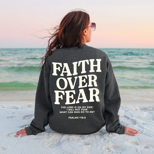 At sunset, a woman sits on the beach wearing sunglasses and a charcoal Faith Over Fear tee or pullover featuring Psalms 118:6. Unisex sizing ensures the perfect fit.