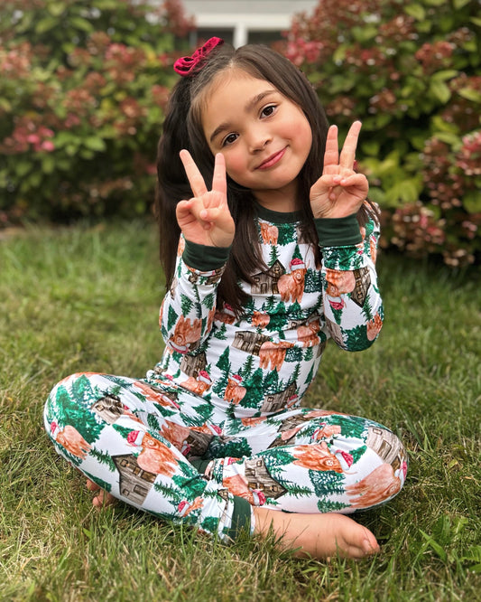 A young girl wears our Santa Cows • 2 Piece Bamboo Set (Updated Fit) pajamas, sitting cross-legged on grass and smiling with peace signs. Pink-flowered bushes add a festive touch to the background.