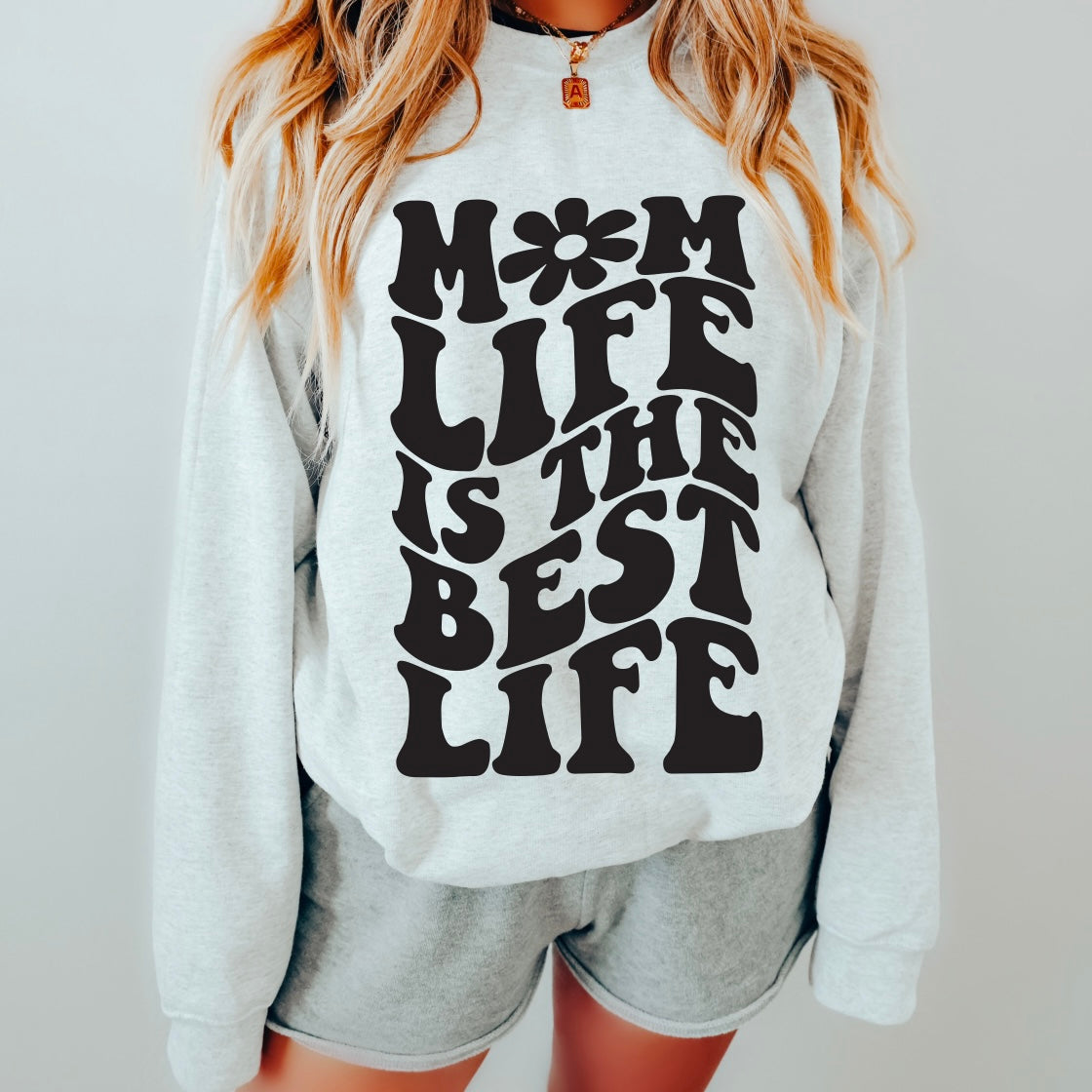 A woman wears the Groovy Mom Life • Ash Pullover, a light gray cotton/polyester sweatshirt with bold black MOM LIFE IS THE BEST LIFE text, paired with matching shorts. Only her torso and part of her legs are visible.