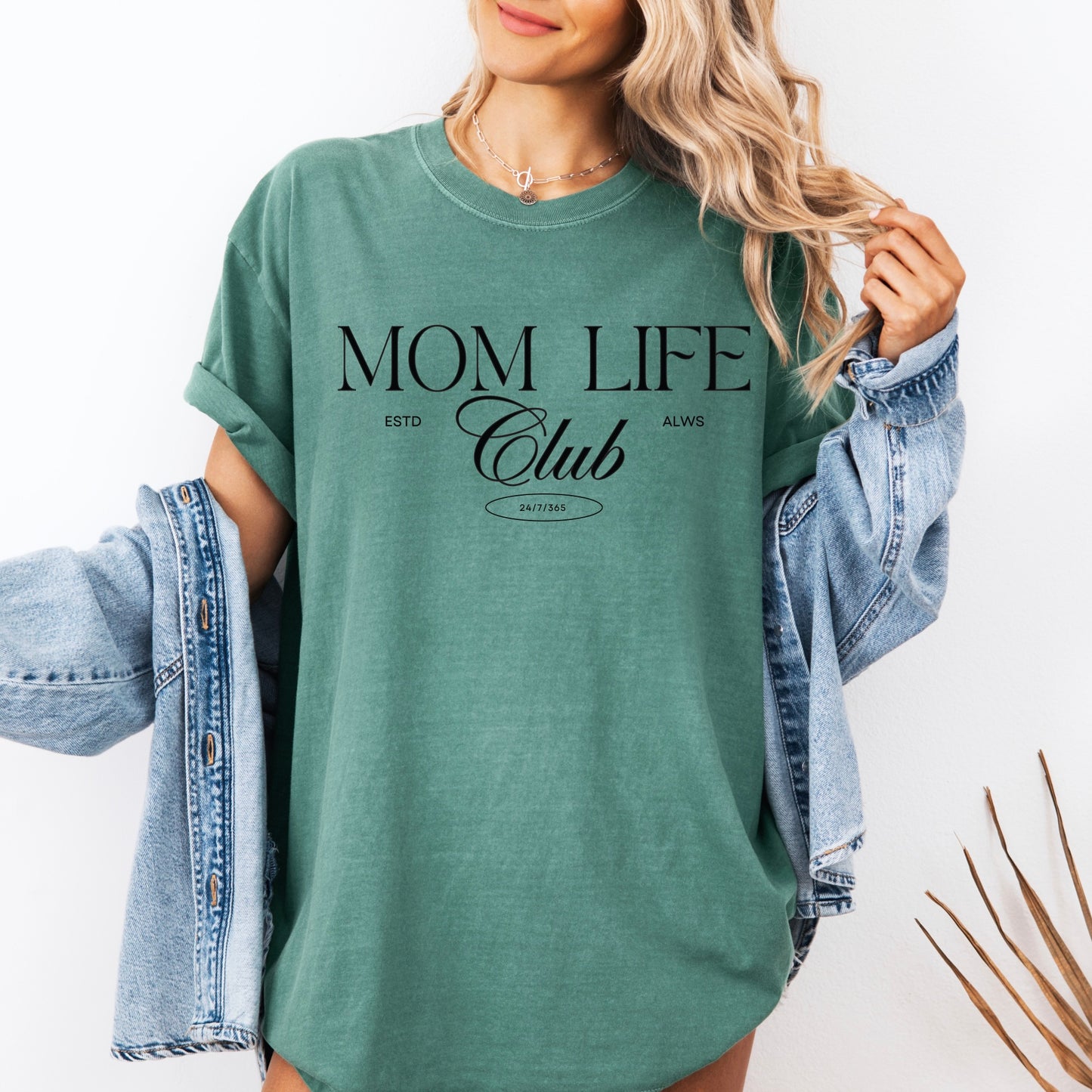A woman in a green Mom Life Club • Comfort Colors Tee stands against a white background, smiling and holding the edges of her shirt with a denim jacket draped over her arms.