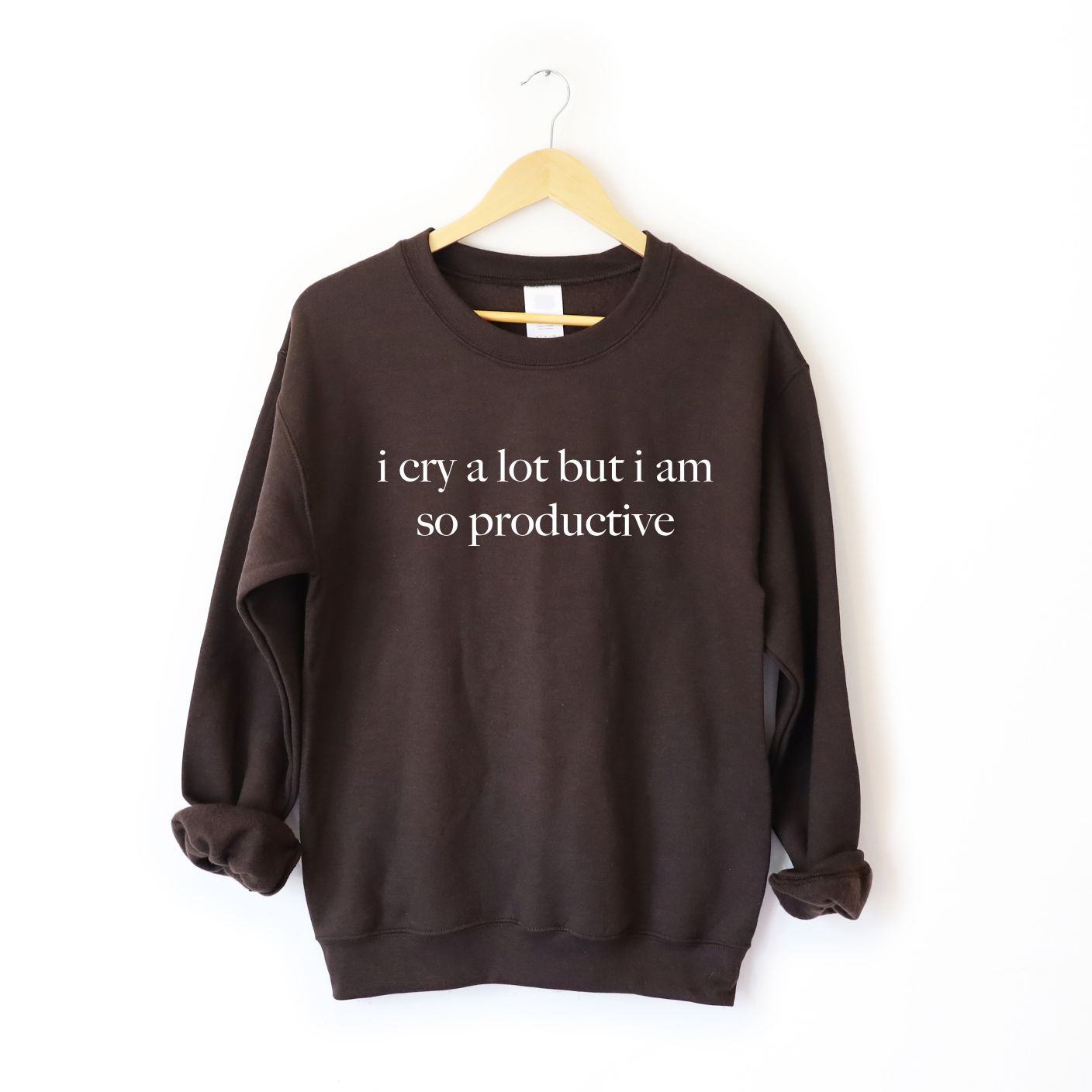 The i cry a lot • Cocoa Pullover is a dark brown unisex sweatshirt with i cry a lot but i am so productive in white, made from a soft cotton-polyester blend. It features a comfy fit and is displayed on a wooden hanger against a plain white background.