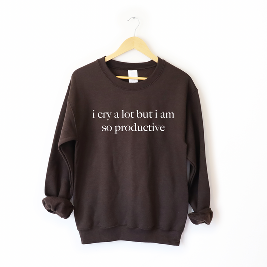 The i cry a lot • Cocoa Pullover is a dark brown unisex sweatshirt with i cry a lot but i am so productive in white, made from a soft cotton-polyester blend. It features a comfy fit and is displayed on a wooden hanger against a plain white background.