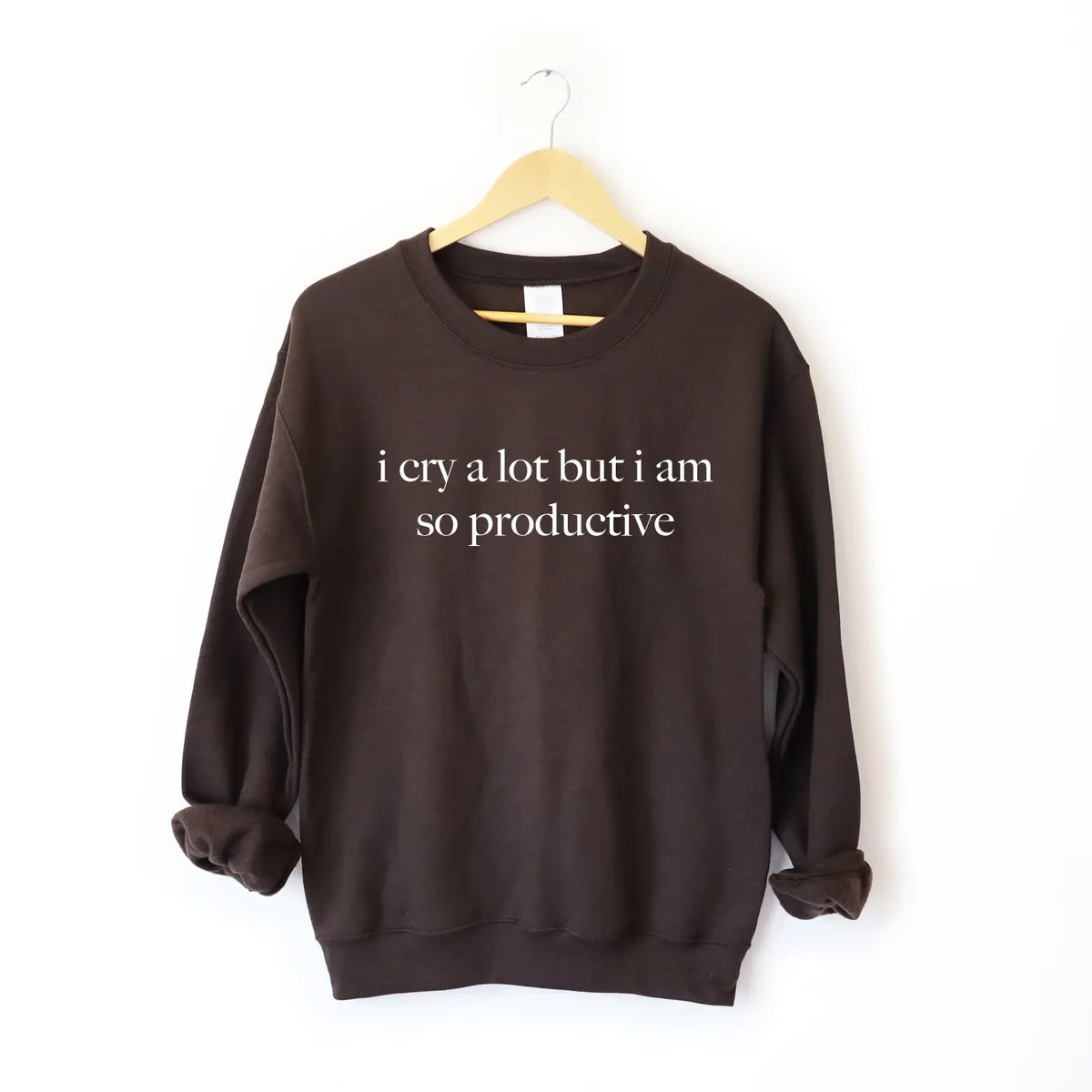 The i cry a lot • Cocoa Pullover is a dark brown unisex sweatshirt with i cry a lot but i am so productive in white, made from a soft cotton-polyester blend. It features a comfy fit and is displayed on a wooden hanger against a plain white background.