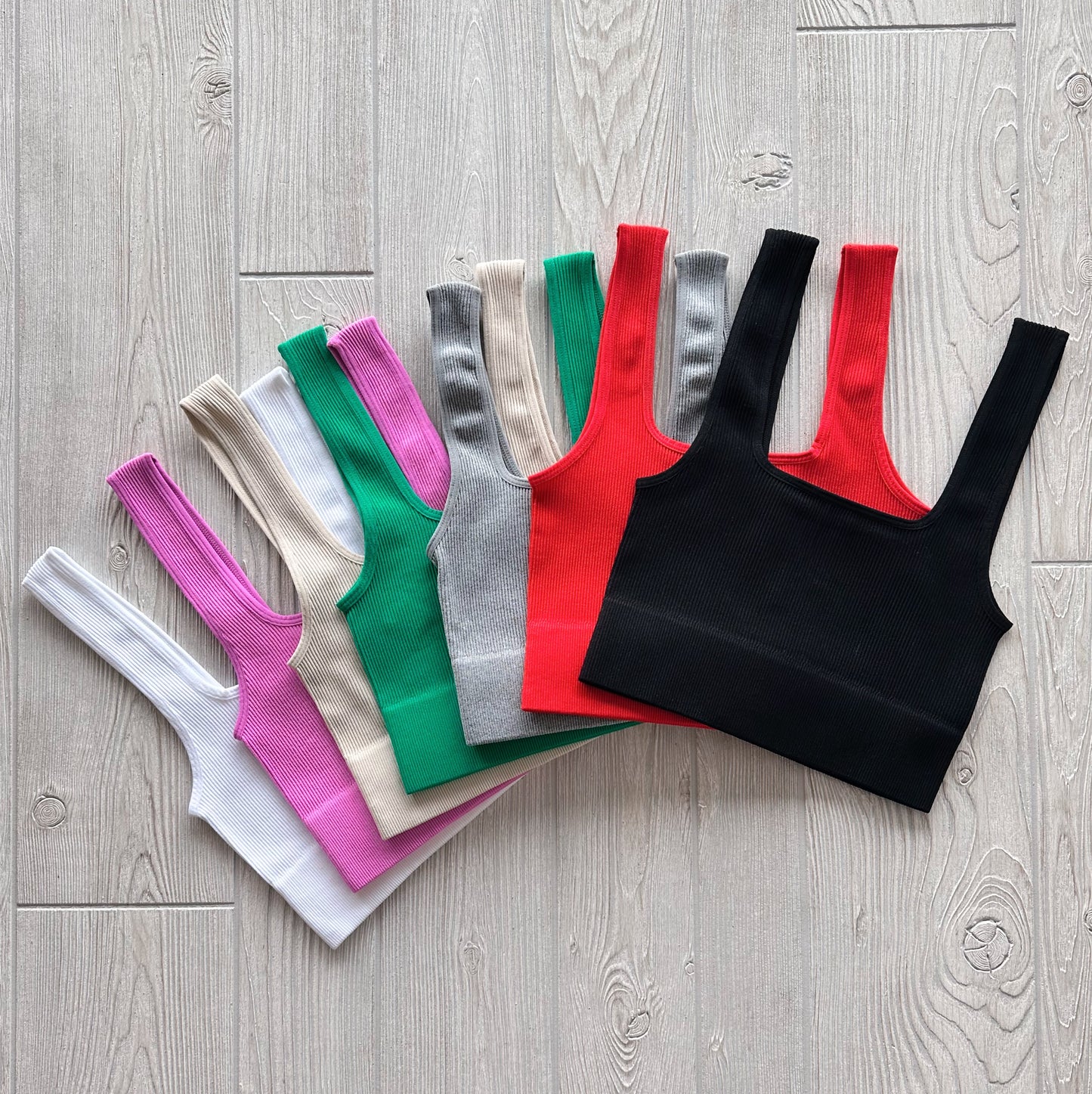 Seven Iris Crop sleeveless ribbed tops in white, lilac, pink, green, beige, gray, and red with black are fanned out on a light wooden floor to highlight their soft stretchy fabric.
