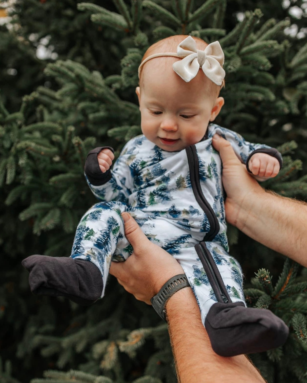 A baby in our Snowy Trees • Bamboo Zippy (Updated Fit) and a beige bow headband is held by an adult before a lush evergreen, looking down with curiosity.