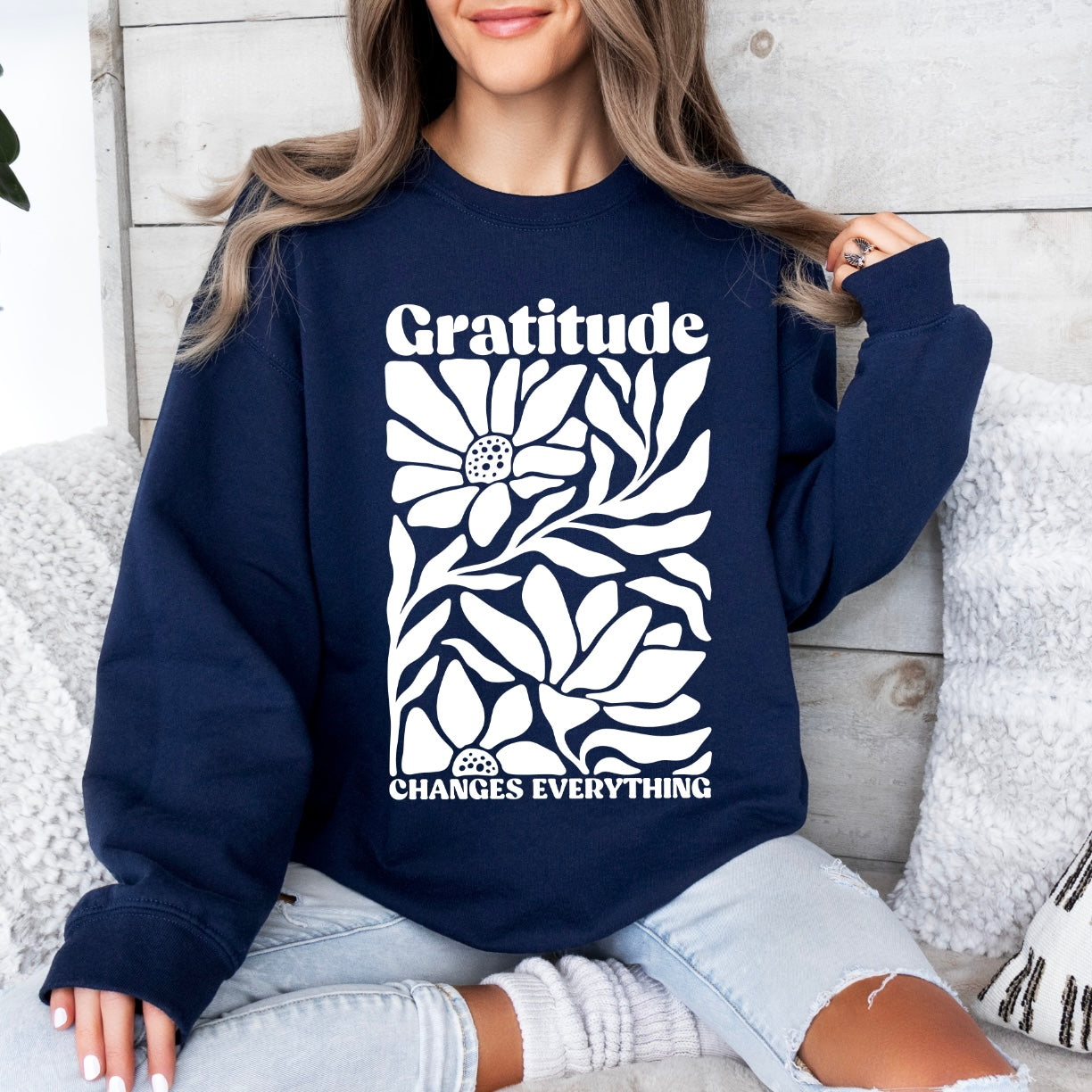 A person wears the Gratitude • Navy Pullover featuring Gratitude Changes Everything and a white floral graphic, relaxing on a couch with light pillows. This unisex sweatshirt is perfect for cozy, casual comfort in cooler weather.