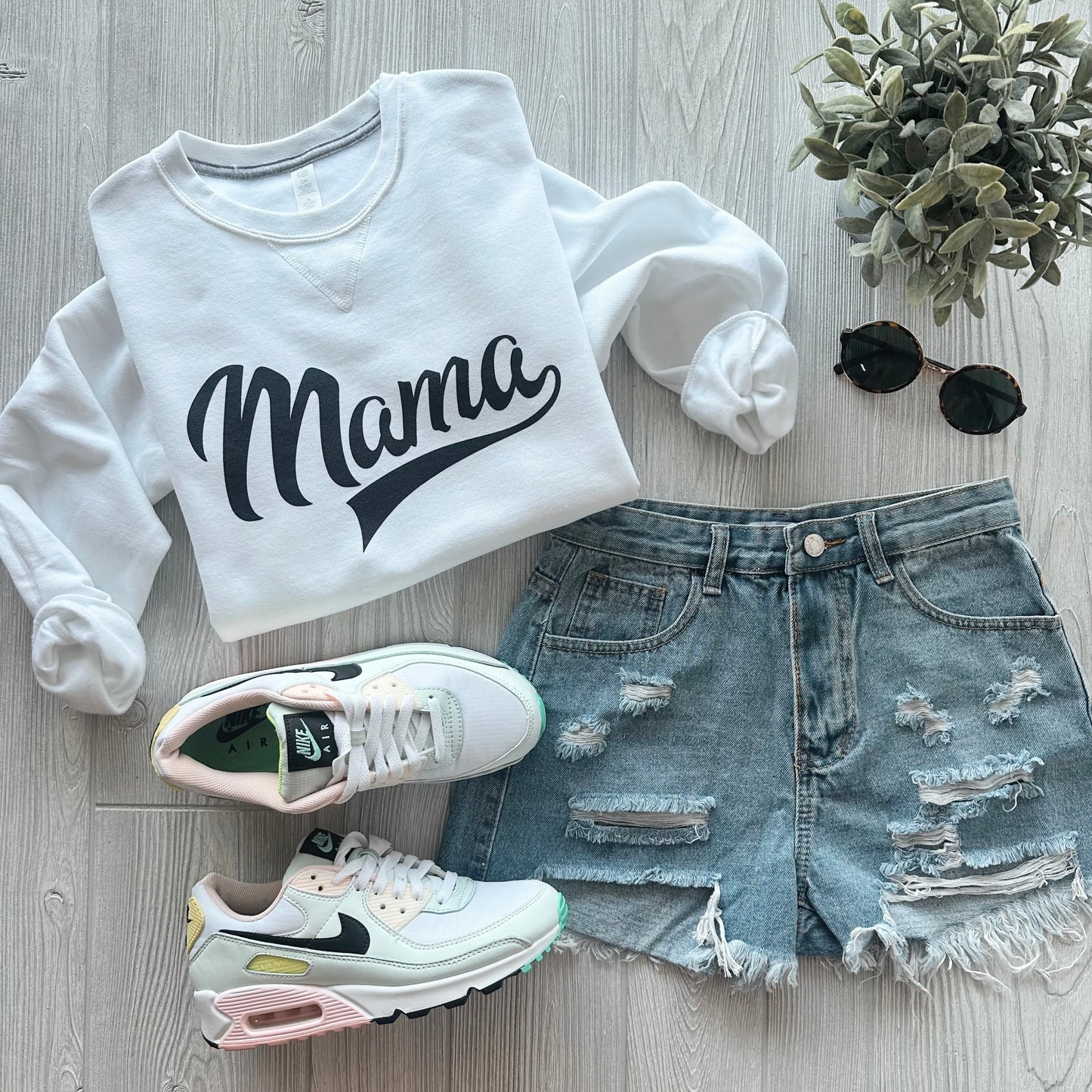 A flat lay of a white Mama Sporty • V-Notch Pullover, distressed denim shorts, pastel-accented white Nike sneakers, black round sunglasses, and a small potted plant on a light wood surface.