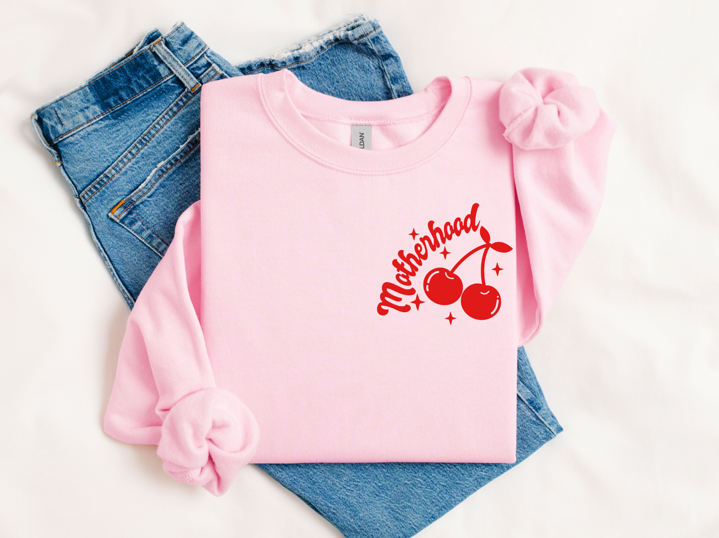 The Motherhood π β’ Pink Pullover, an oversized light pink sweatshirt with red cherries and Motherhood in red script, is styled with light blue jeans and two matching pink scrunchies on a white background.