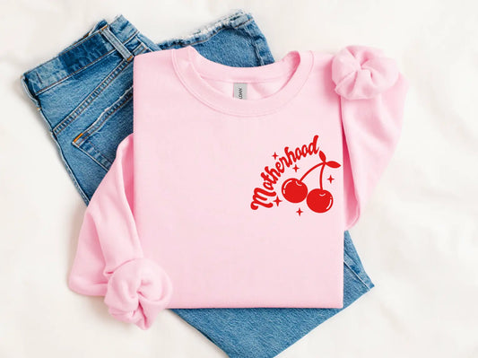 The Motherhood 🍒 • Pink Pullover, an oversized light pink sweatshirt with red cherries and Motherhood in red script, is styled with light blue jeans and two matching pink scrunchies on a white background.