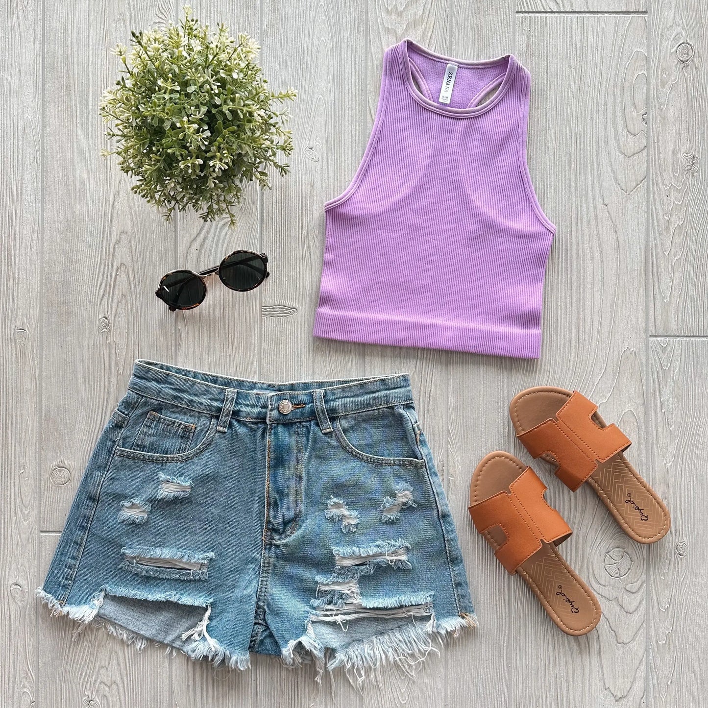 Flat lay of a summer outfit: Sophia High-Neck Crop in lavender, distressed denim shorts, tan slide sandals, black round sunglasses, and a small green potted plant on a light wood floor.
