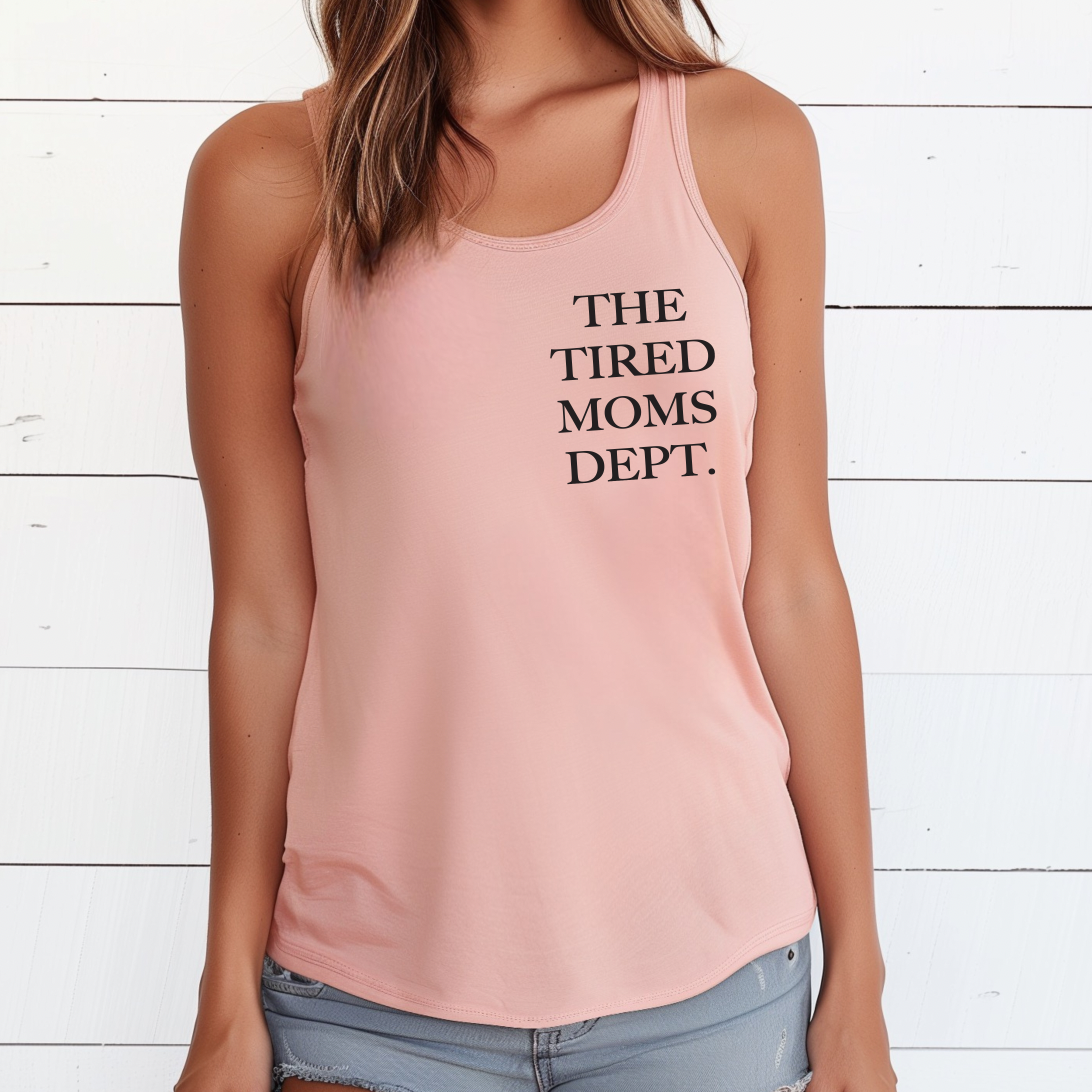 A woman stands before a white wooden wall wearing the TTMD • Desert Pink Tank, a light pink racerback top with THE TIRED MOMS DEPT. printed in black on the front.
