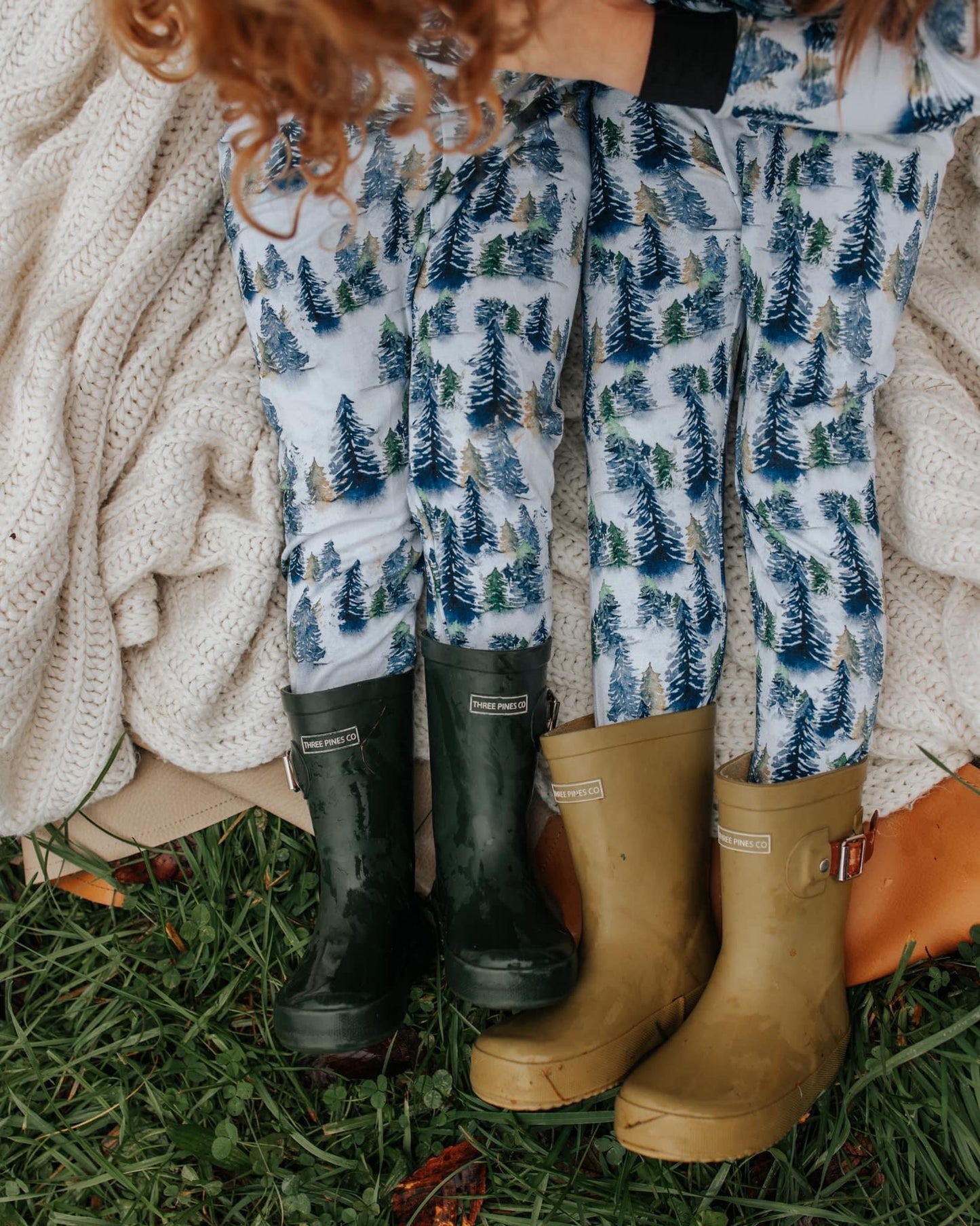 Two children lounge on a cream knit blanket in Snowy Trees • 2 Piece Bamboo Set (Updated Fit) pajamas—eco-friendly and gentle for sensitive skin. Green and tan rubber boots peek out as they sit on grass; only their legs and part of ones hair are visible.