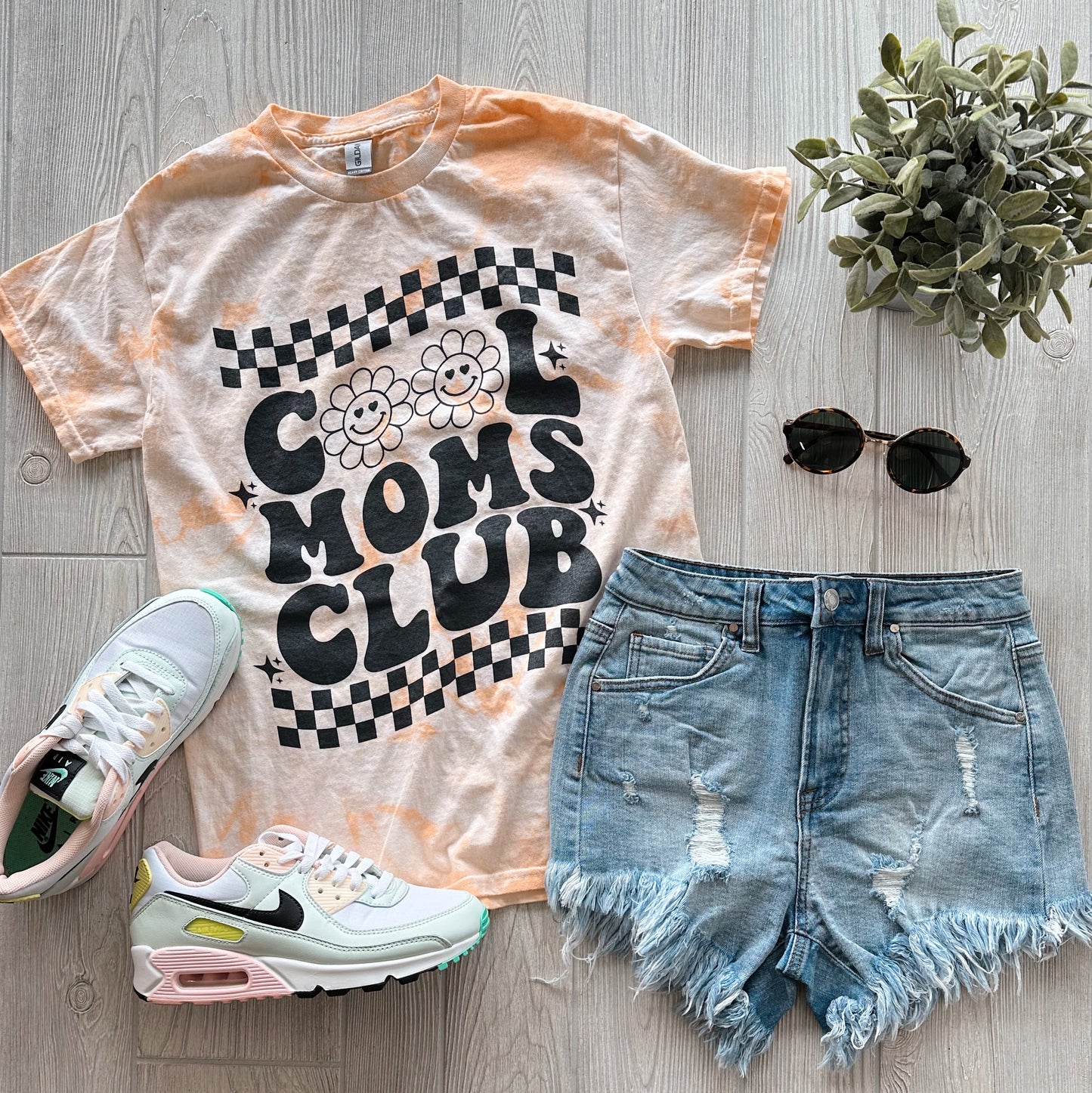 Flat lay of a casual outfit with the Cool Moms • Mango Tie Dye unisex tee, distressed denim shorts, Nike sneakers, round sunglasses, and a small potted plant on a light wooden floor.