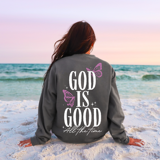 A woman with long hair sits on a white sandy beach at sunset, wearing an Asphalt God is Good Tee or Pullover featuring pink butterflies and “God is Good All the Time” in white script on the back.