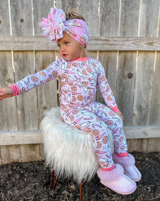 A young child from the Cozy Collection wears the Milk & Cookies • 2 Piece Bamboo Set (Updated Fit) with cookie and milk patterns, a pink bow headband, and slippers, sitting on a furry white stool by a wooden fence, reaching out to the side.