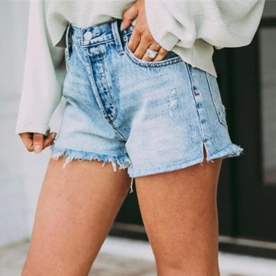 A person wears Myra Denim Shorts • Medium Wash—high-waisted womens jean shorts with frayed hems—paired with a light long-sleeve top. The distressed denim adds trendiness, and the blurred background highlights the outfit.