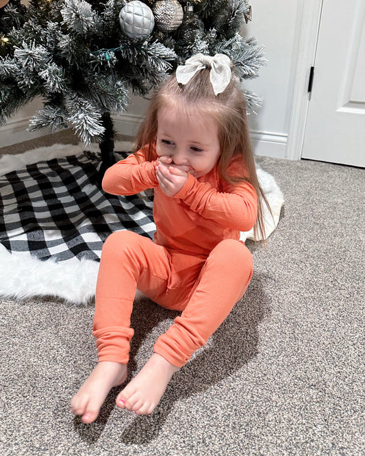 A young girl in the Coral Ribbed Solid • 2 Piece Bamboo Set sits on a carpet by a Christmas tree with a buffalo plaid skirt and white, silver, and gray ornaments, covering her mouth with both hands and smiling. Perfect for sensitive skin.