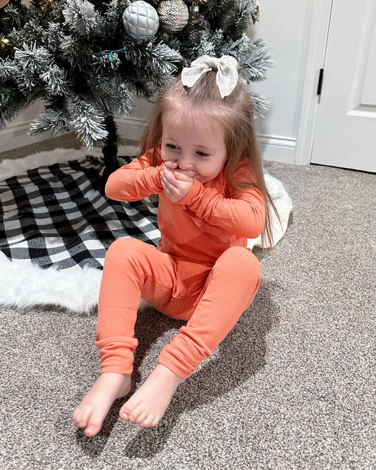 A young girl in the Coral Ribbed Solid • 2 Piece Bamboo Set sits on a carpet by a Christmas tree with a buffalo plaid skirt and white, silver, and gray ornaments, covering her mouth with both hands and smiling. Perfect for sensitive skin.