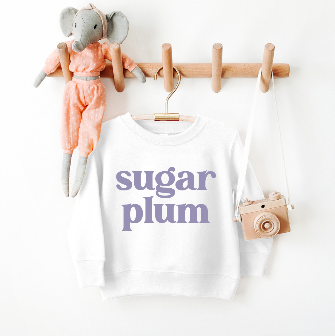 The Sugar Plum • Kids Sweatshirt, a white pullover with sugar plum in bold purple letters, hangs on a wooden hook rack in toddler/youth sizes beside a pink plush elephant doll and a toy camera with a white strap.