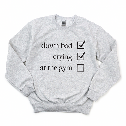 The Not at the Gym • Ash Pullover is a light gray cotton/polyester unisex sweatshirt with down bad and crying marked and at the gym unmarked in black text on the front.
