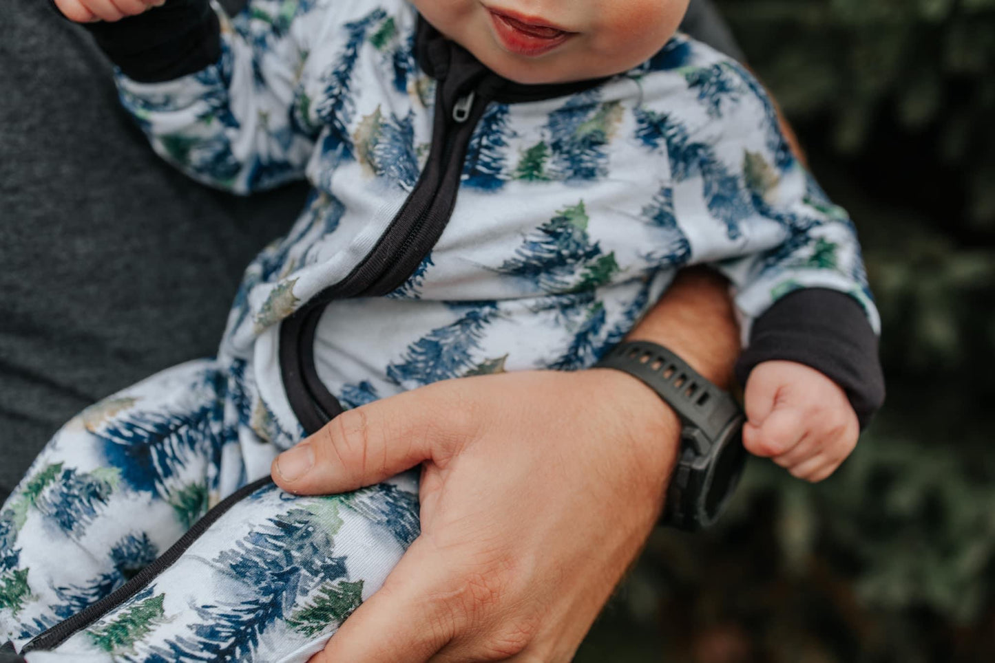 An adult cradles a baby in the Snowy Trees • Bamboo Zippy (Updated Fit) onesie. The babys face is partly shown, with the adults hand and watch in focus. Trees from the cozy collection create a soft, blurred background.