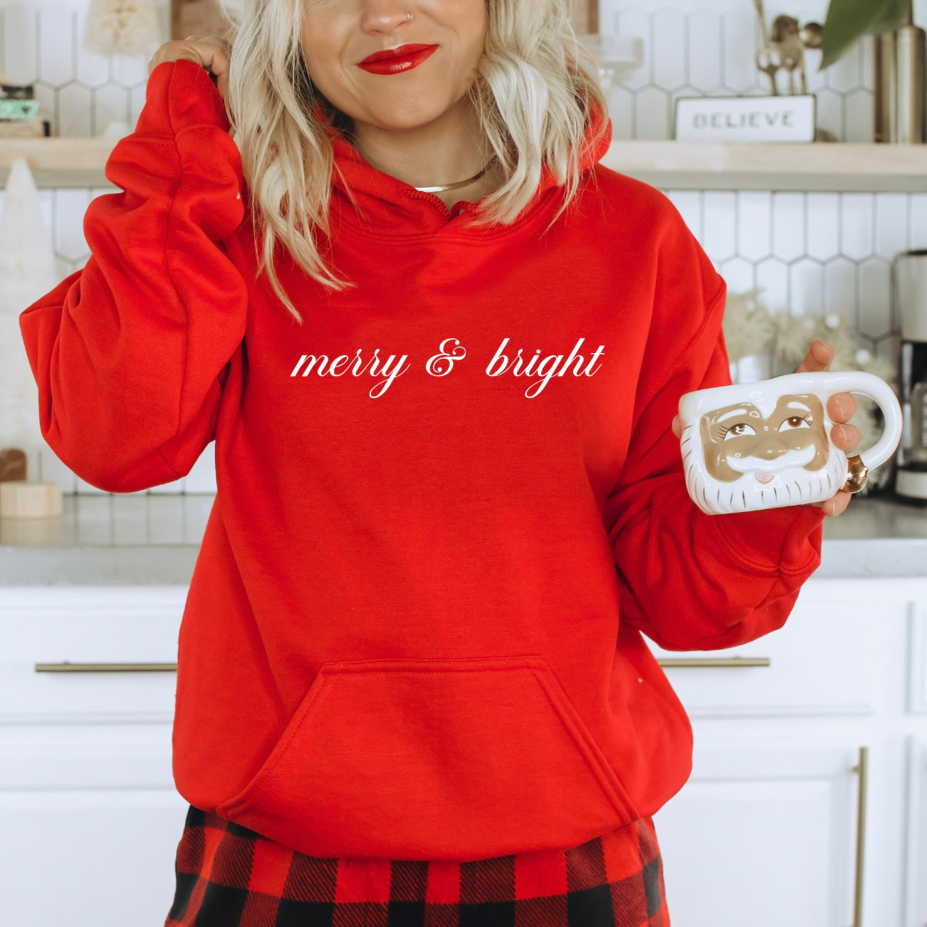 A woman in a cozy merry & bright Red Hoodie and plaid pants holds a Santa mug, standing in a festive kitchen decorated for the holidays. This unisex hoodie is perfect for celebrating the season.