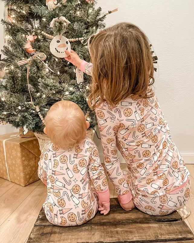 Two children wearing our Milk & Cookies • 2 Piece Bamboo Set (Updated Fit) sit on a wooden floor, facing away as they decorate a Christmas tree together. Wrapped gifts are visible under the tree.