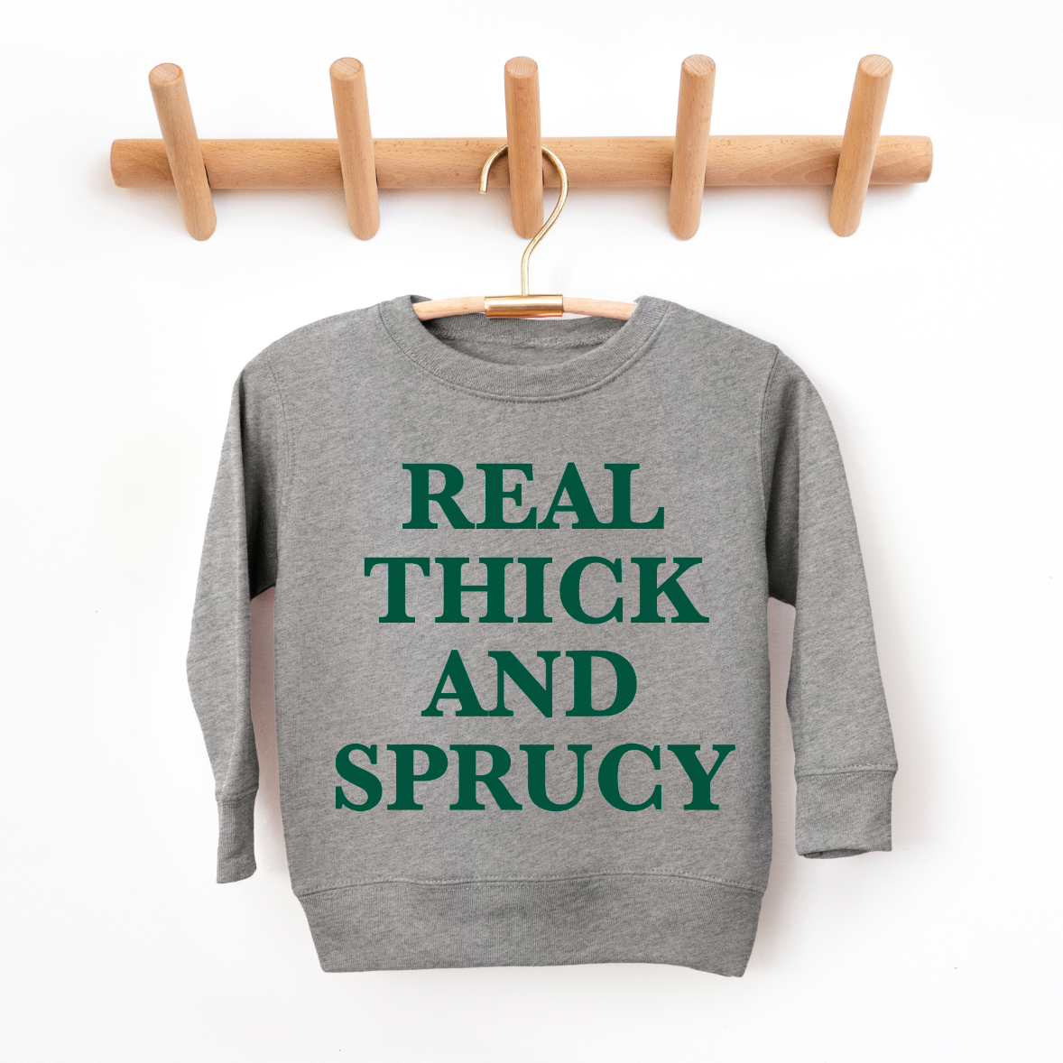 The THICK & SPRUCY • Kids Sweatshirt, a gray unisex top with REAL THICK AND SPRUCY in bold green letters, is shown on a wooden hanger and wall rack against a white background. Available in toddler and youth sizes.