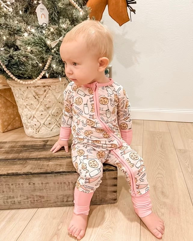 A baby in our Milk & Cookies • Bamboo Zippy (Updated Fit)—pink and white pajamas with cookie and milk designs—sits on a wooden box by a decorated Christmas tree indoors, looking to the left.
