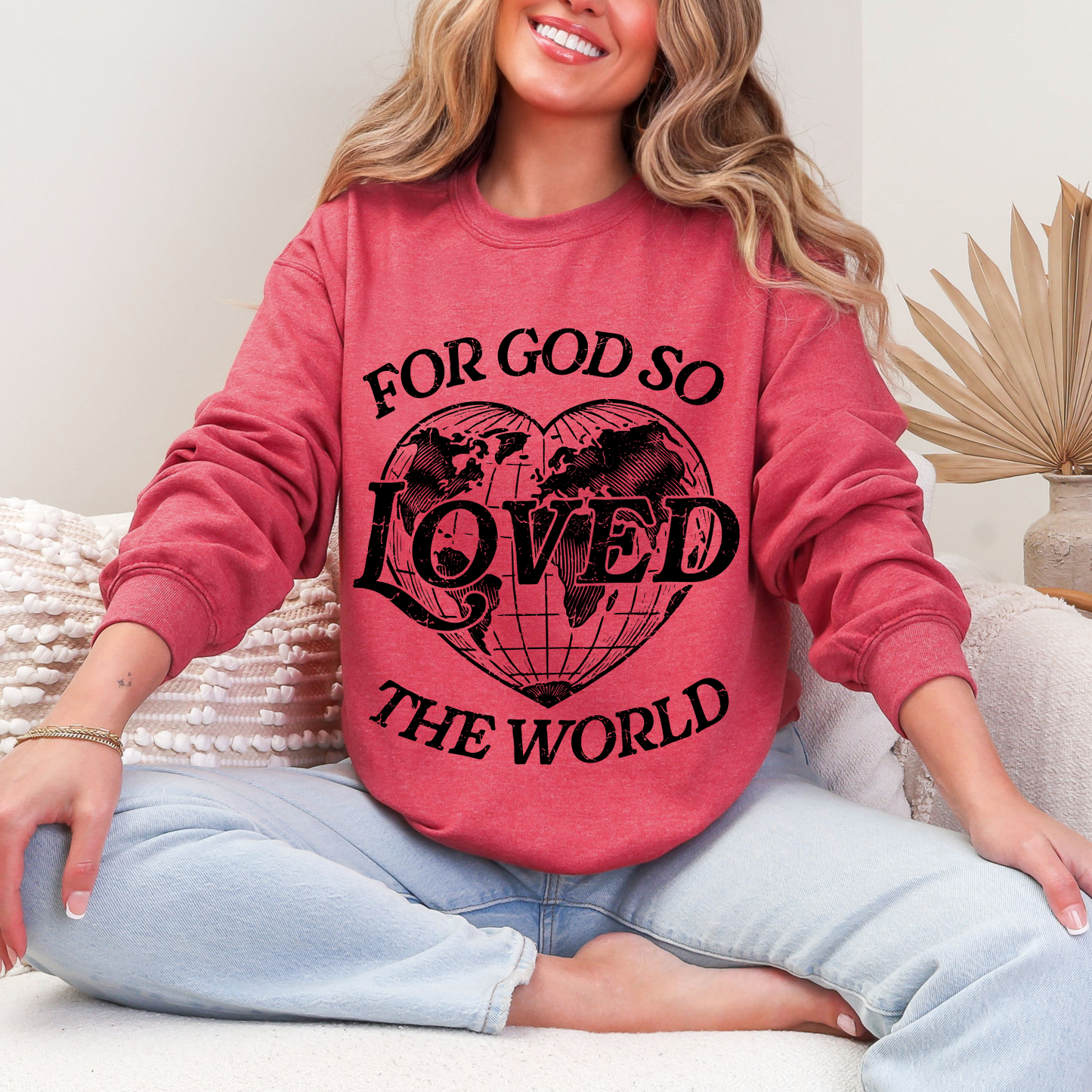 A smiling woman sits cross-legged on a couch, wearing the Loved the World • Red Pullover—a heather red fleece sweatshirt featuring “For God so loved the world” with a heart and world map graphic.
