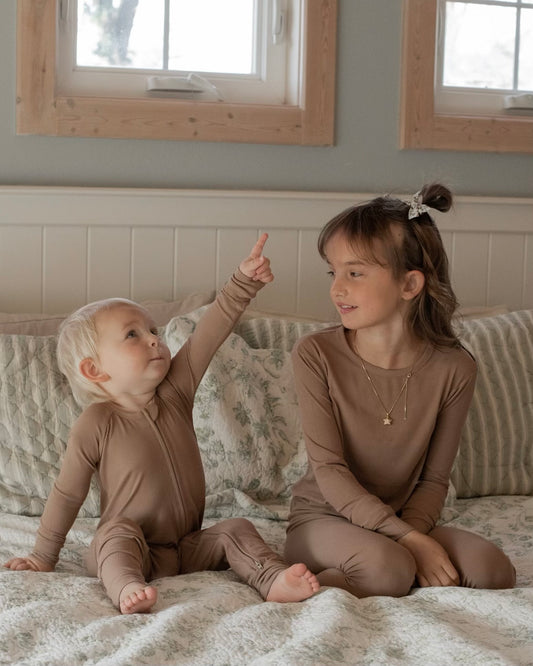 A young child in Chestnut Ribbed Solid • Bamboo Zippy (Updated Fit) pajamas sits on a bed pointing upward, next to an older child in matching pajamas, both on patterned bedding in a cozy bedroom.