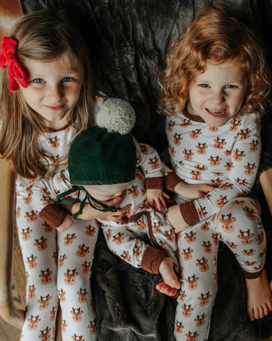 Three young children sit together on a dark blanket, all in cozy Rudolph • 2 Piece Bamboo Set pajamas with reindeer prints. One wears a red bow, another has curly hair, and the youngest sports a green hat with a pom-pom.