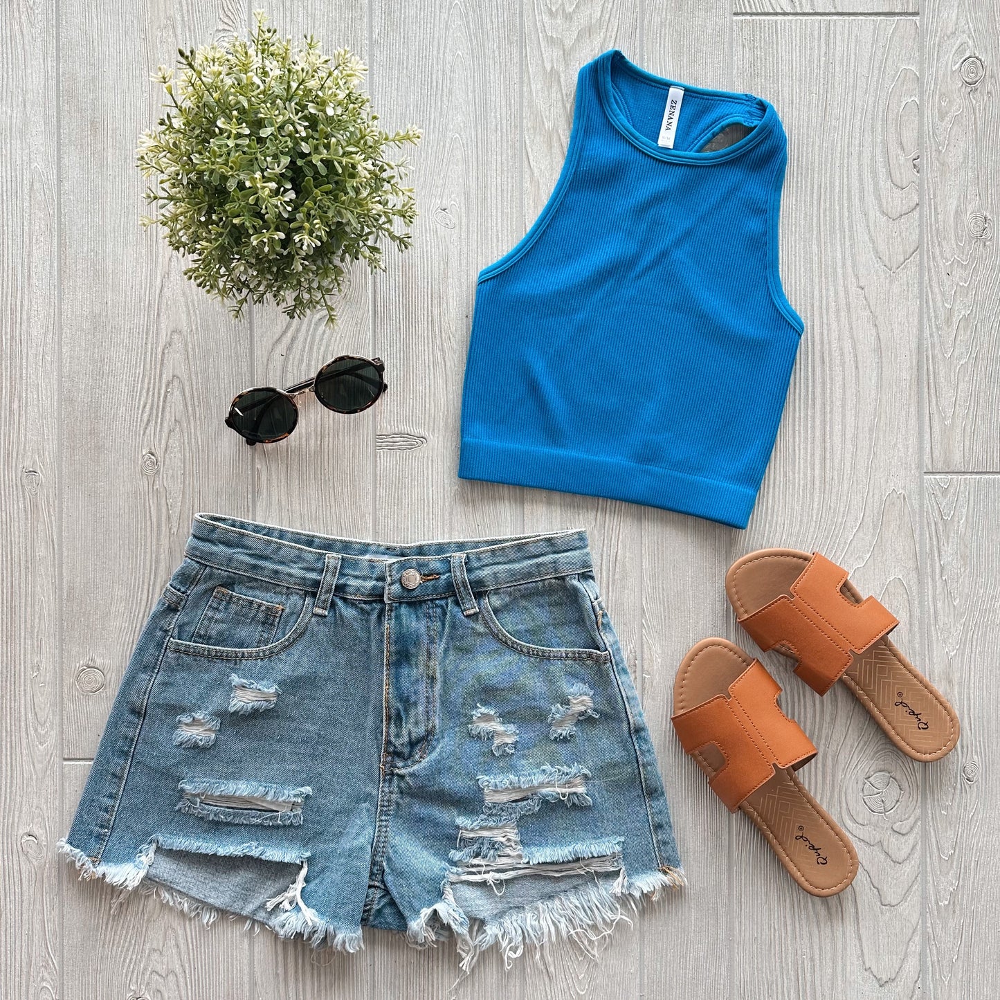 Flat lay of a summer outfit: Sophia High-Neck Crop in Aqua, distressed denim shorts, tan slide sandals, black round sunglasses, and a small potted plant on a light wooden floor—ideal for casual workouts or relaxed days out.