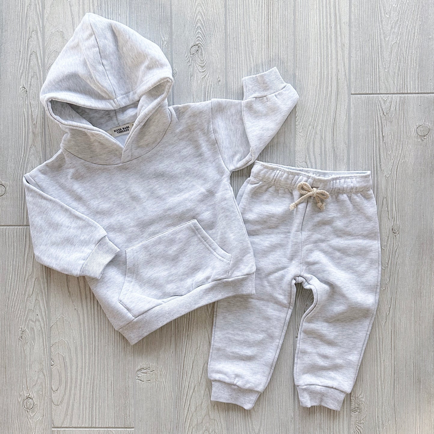 The Ash • Sweatpant & Hoodie Set is a light gray organic two-piece for babies and toddlers, with a cozy fleece hoodie (front pocket) and matching cuffed joggers, both laid flat on a light wood floor.