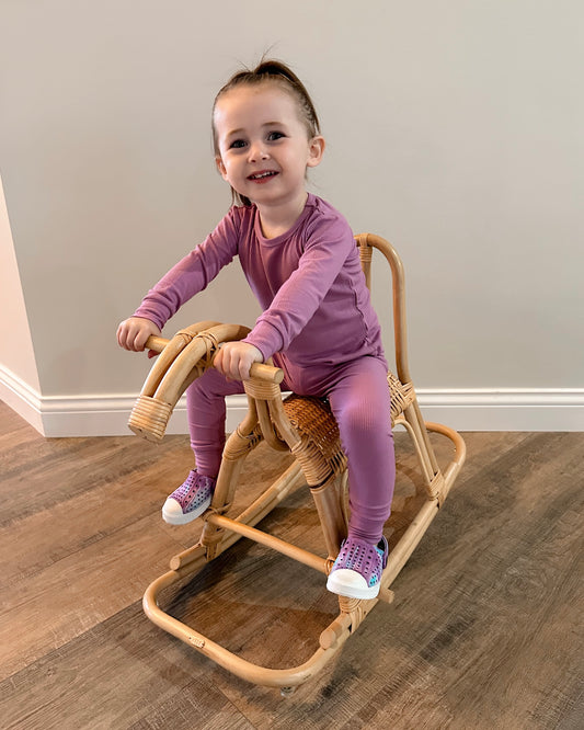 A young child in the Lilac Ribbed Solid • 2 Piece Bamboo Set (Updated Fit) smiles while sitting on a wicker rocking horse indoors.